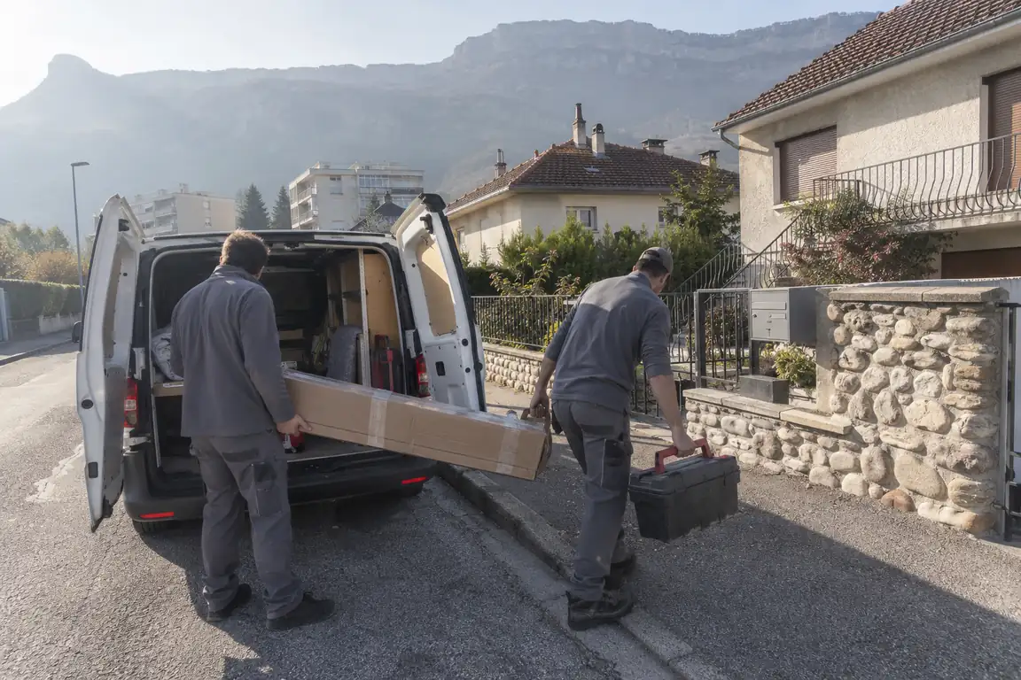 Deux poseurs déchargeant un carton de leur utilitaire dans une rue résidentielle, falaises de la Chartreuse au fond