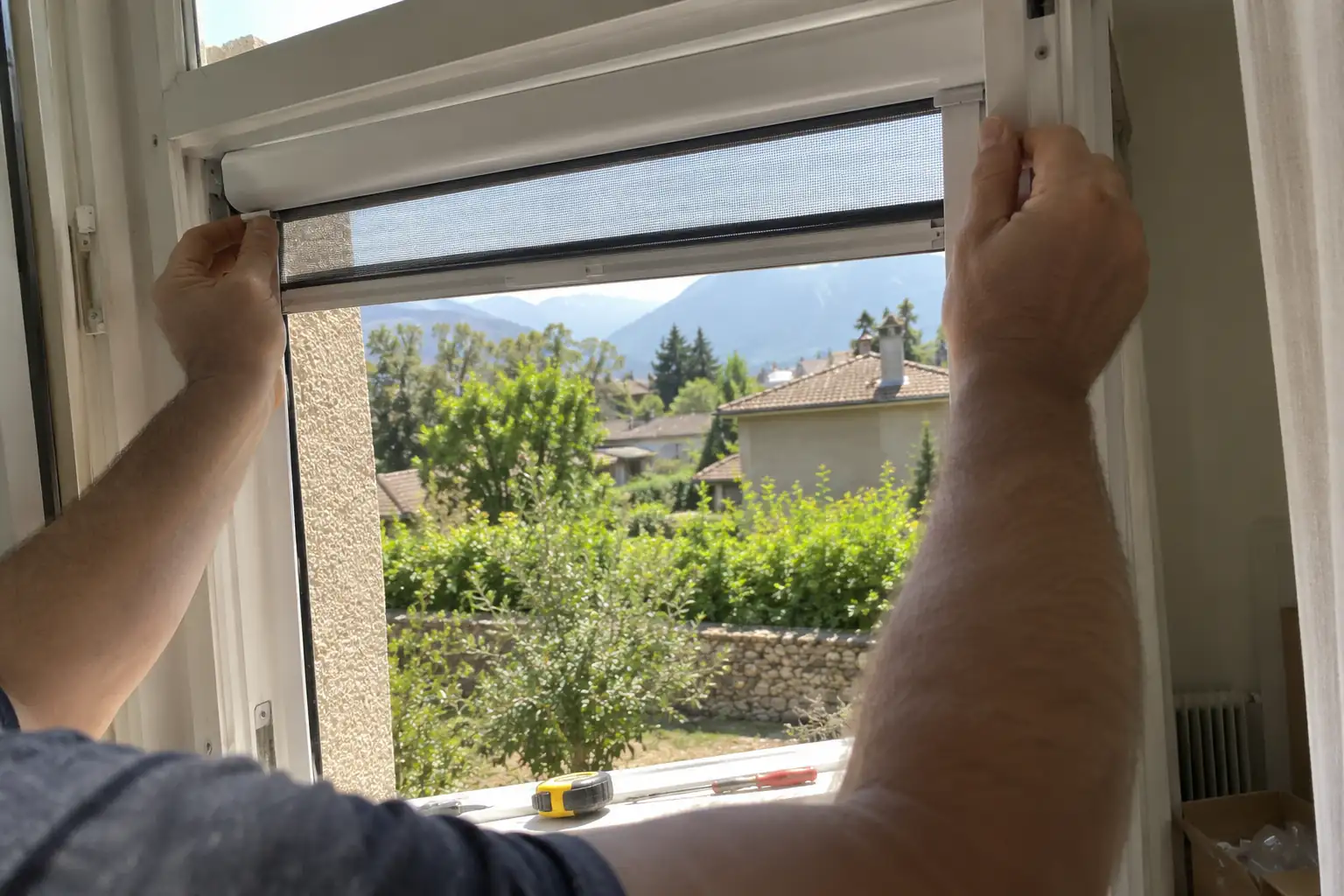 Mains ajustant le cadre d’une moustiquaire enroulable dans l’embrasure d’une fenêtre ouverte sur un jardin