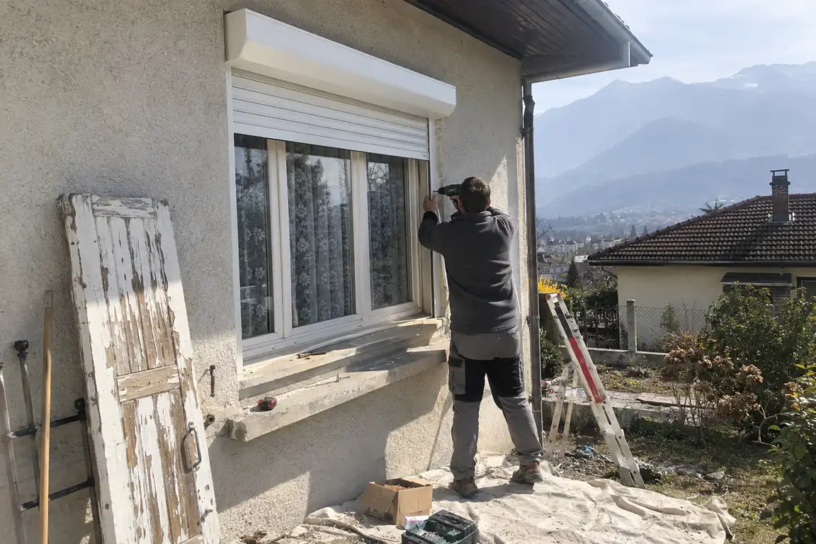 Poseur fixant un volet roulant sous le linteau d’une fenêtre, ancien volet battant en bois appuyé contre le mur