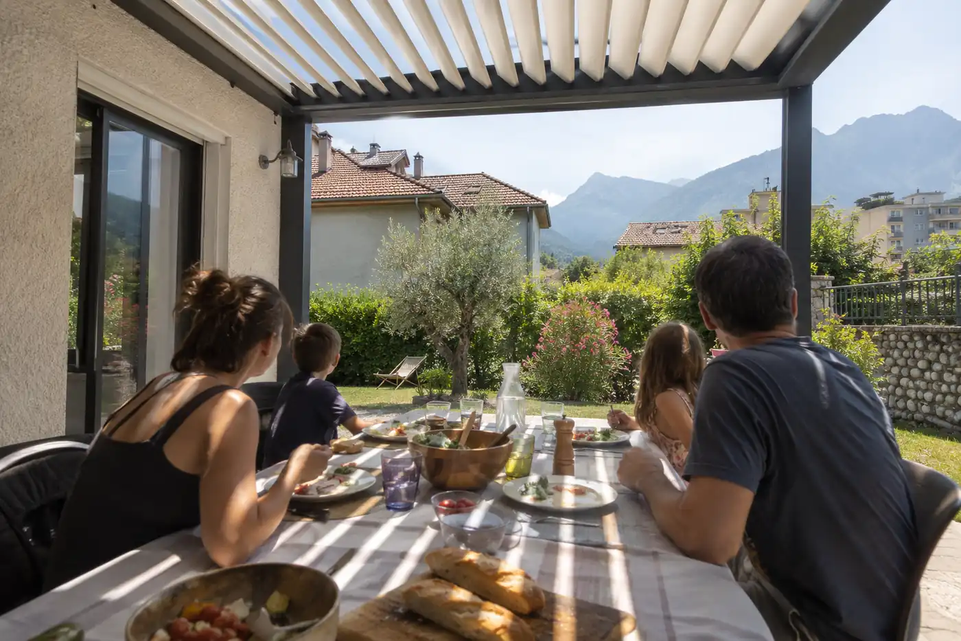 Famille attablée pour un repas sous une pergola aux lames entrouvertes, lumière filtrée sur la nappe