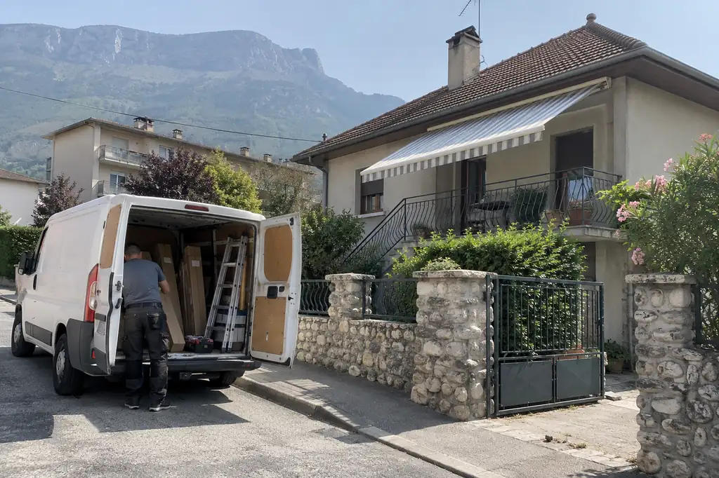 Utilitaire de pose garé portes ouvertes devant une maison au muret de galets, store banne déployé à l’étage